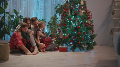 A-family-with-two-children-sit-together-at-the-Christmas-tree-and-cuddle.-Love-and-family-happiness-on-Christmas-Eve.-Father-and-two-sons-cuddle-and-look-at-Christmas-tree-in-their-living-room.-High-quality-4k-footage